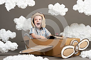 The boy plays in an airplane made of cardboard box and dreams of becoming a pilot, clouds from cotton wool on a gray background, r