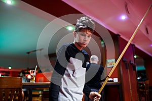 Boy plays billiard or pool in club. Young Kid learns to play snooker. Boy with billiard cue strikes the ball on table.