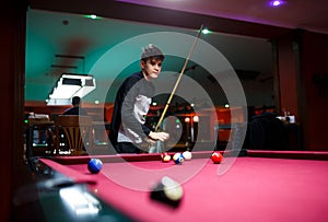 Boy plays billiard or pool in club. Young Kid learns to play snooker. Boy with billiard cue strikes the ball on table.