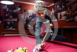 Boy plays billiard or pool in club. Young Kid learns to play snooker. Boy with billiard cue strikes the ball on table.