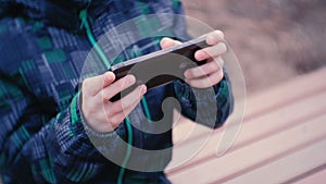 Boy plays a game on his mobile phone sitting in the Park on a bench. Close-up boy`s hands.