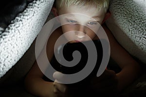 Boy plays games on a smartphone at night under a blanket on the floor