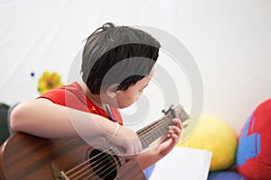 The boy plays the guitar with his left hand, a song by himself, on the sofa, in the bedroom, reading the sheet music