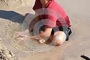 A boy plays in a puddle on the beach. The child sits in a puddle in the sand. Happy funny summer.