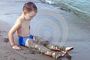 A boy plays on the seashore with sand. The child laid sand on his feet.
