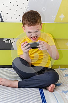 The boy plays on a smartphone while sitting in his room on the bed.