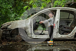 A boy pretends to be a driver while standing next to a scrapped car