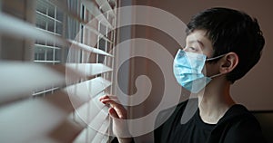 A boy with a protection mask looks out the window. Coronavirus, Covid-19.