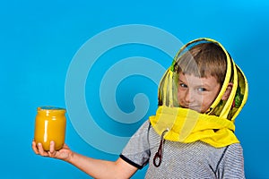 A boy in a protective mask from bees holds in his hand a jar of honey on a blue background