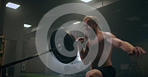 The boy pushes the barbell in the gym with one hand. An athlete without a T-shirt in a dark room lifts the barbell while