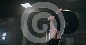 The boy pushes the barbell in the gym with one hand. An athlete without a T-shirt in a dark room lifts the barbell while