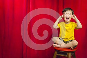 Boy Puts Fingers In Ears on Stool in Front of Theater Curtain