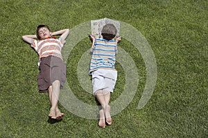 Boy Reading Besides Asleep Brother On Grass