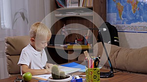 A boy is reading a book at home at his desk, a backpack is in the background.