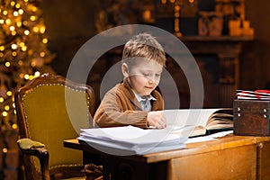 Boy reads a magic book while sitting at the table. Home interior with Christmas tree and fireplace. Traditional