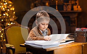 Boy reads a magic book while sitting at the table. Home interior with Christmas tree and fireplace. Traditional