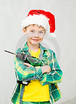 Boy in a red christmas hat with a drill in hands looking at camera