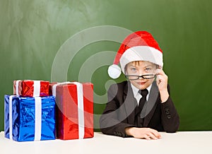 Boy in red christmas hat with gift boxes near empty green blackboard