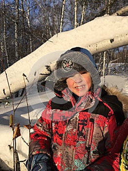 A boy in a red jacket and a hat with earflaps on the background of a fallen birch trunk in the snow