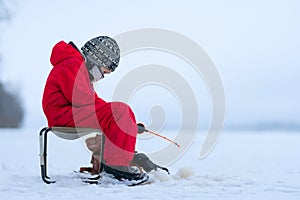 A boy in red jumpsuit on a frozen snowy lake. Fishing rod in the hands of a boy. Winter fishing in ice hole.