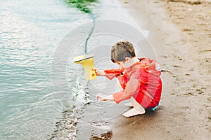 Boy in a red raincoat pours water out of yellow rubber boot into the lake. Child playing with water at pond. Kid having