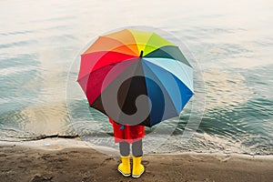 Boy in a red raincoat and yellow rubber boots holds rainbow umbrella standing at beach. Child with colourful umbrella