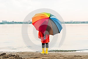 Boy in a red raincoat and yellow rubber boots holds rainbow umbrella standing at beach. Child with colourful umbrella