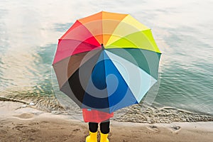 Boy in a red raincoat and yellow rubber boots holds rainbow umbrella standing at beach. Child with colourful umbrella