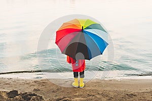 Boy in a red raincoat and yellow rubber boots holds rainbow umbrella standing at beach. Child with colourful umbrella