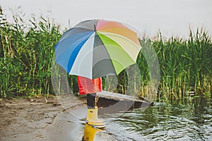 Boy in a red raincoat and yellow rubber boots stands at river bank and holding rainbow umbrella. School kid standing