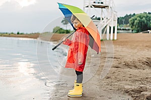 Boy in a red raincoat and yellow rubber boots stands at river bank and holding rainbow umbrella. School kid standing