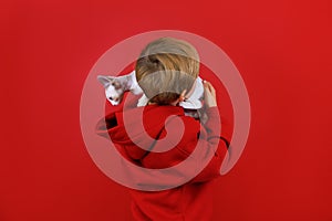 a boy in a red suit stands on a red background, turned his back, holds a small white kitten on his shoulder, which looks around