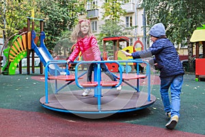 boy rolls girl in pink jacket on carousel in yard at playground outdoors