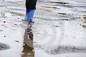 Boy in rubber blue rainboots jumping to dirty puddle