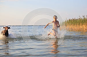 A boy runs with the dog in the lake, splashing the water around. Playful, happy childhood moments. Beautiful sunny summer day.