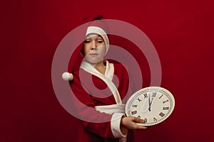 A boy in a Santa costume holds a white watch on a red background.