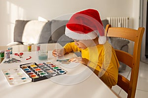 Boy in santa hat drawing at home, occupation and activity for small children before christmas, watercolors for kids