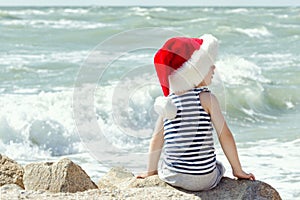 Boy in santa hat sitting on a rock. Sea shore. Back view