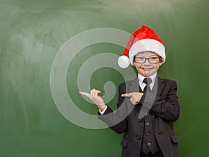 Boy in Santa red hat points on empty green chalkboard