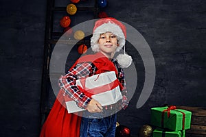 A boy in Santa`s hat holds gift box and red bag.