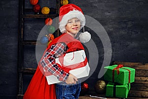 A boy in Santa`s hat holds gift box and red bag.