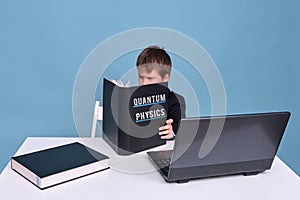 A boy in a school suit with a textbook on quantum physics during distance learning, copy space on a blue studio background