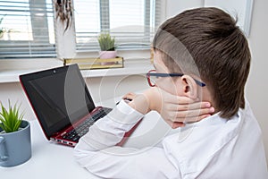 A boy, schoolboy in white formal shirt sitting by the table, desk with laptop and doing homework at home, e-learning, distant