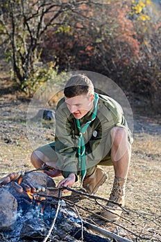 Boy Scout Cooking Sausages on Sticks over Campfire