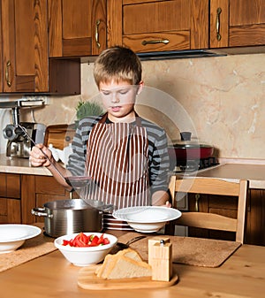 Boy serving Borshch, traditional Russian and Ukrainian soup. Pouring soup into a plate with ladle from pan in kitchen.