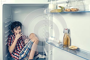 A boy in a shirt and shorts eating a chocolate bar inside a fridge with food