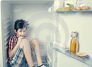 A boy in a shirt and shorts eating a chocolate bar inside an open fridge with food