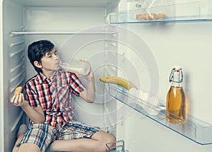 A boy in a shirt and shorts eating a croissant and drink milk inside a fridge with food and product. Close-up