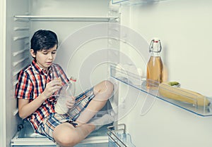 A boy in a shirt and shorts eating a croissant and drink milk inside a fridge with food and product. Close-up