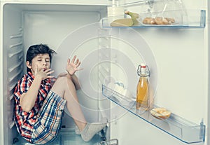 A boy in a shirt and shorts licking one`s fingers inside an open fridge with food remains